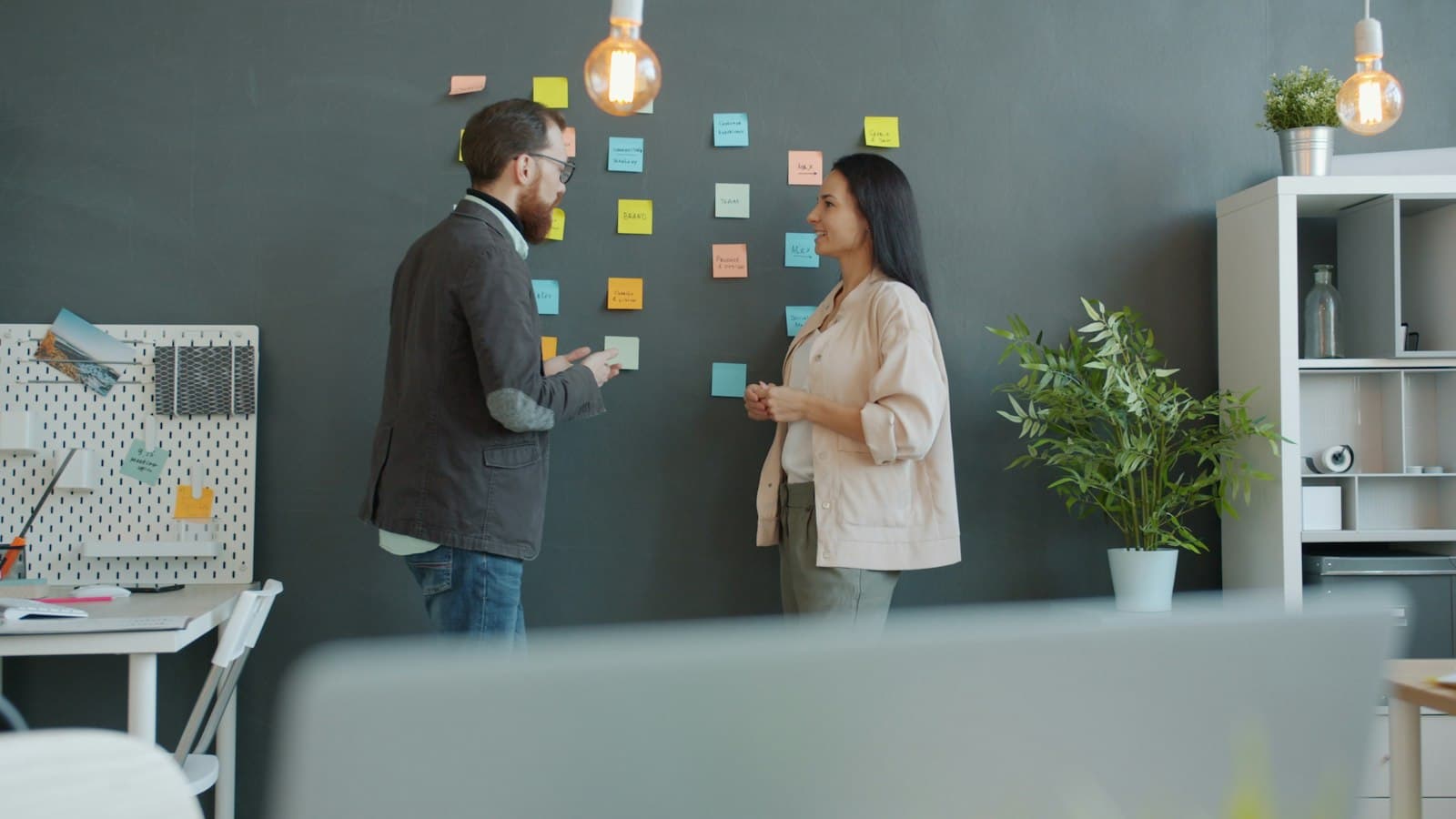 Two colleagues discussing ideas on sticky notes stuck to a wall in a modern office